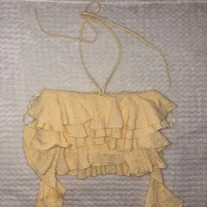 Gul volang topp från Zara - Supersöt gul topp från Zara med flera lager volanger och tunna knytband i nacken. Toppen är ärmlös och har en croppad passform, perfekt för att skapa en somrig och trendig look. Materialet är lätt och har en skrynklig struktur.