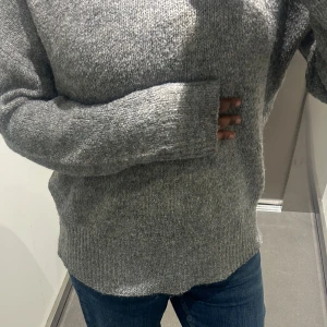 Grå stickad tröja från 157, storlek M - Mysig grå stickad tröja från 157 i storlek M. Tröjan har en klassisk rak passform, långa ärmar och ribbade muddar vid ärmslut och nederkant. Perfekt för lager-på-lager och chill dagar.