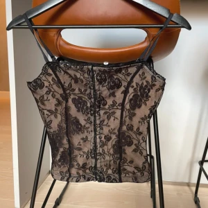 Svart spetskorsett från Collusion 34 - Säljer en snygg svart korsett från Collusion med blommigt spetsmönster och tunna axelband. Toppen har en beige bas och svarta detaljer som ger en cool kontrast. Perfekt för dig som vill sticka ut med en trendig och feminin look.