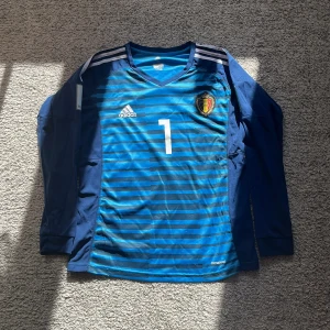 Belgien Courtois målvaktströja VM 2018 Storlek s  - Säljer en blå långärmad Belgien målvaktströja från VM 2018 med Courtois och nummer 1 på ryggen. Adidas ClimaCool-material, randigt mönster framtill, officiellt FIFA World Cup-märke på ärmen och Belgien-emblem på bröstet. Perfekt för fotbollsfans! Pris kan diskuteras 