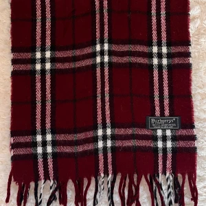 Rutig vinröd halsduk från Burberrys - Snygg halsduk från Burberrys i klassiskt rutig design med vinröd, svart, vit och rosa färg. Gjord av 100% lammull, mjuk och varm med fransar i ändarna. Perfekt för att ge din outfit en cool och tidlös vibe. Priset kan diskuteras vid köp.