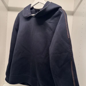 Svart hoodie från Emporio Armani - Svart hoodie från Emporio Armani med broderad logga längs ena ärmen. Tröjan har huva med tryckknappar och sportig känsla. Mjuk och skön, perfekt för chill dagar. Klassisk passform och stilren design.