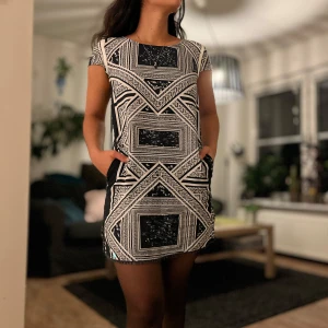 Kortklänning med fickor - Säljer en svartvit kortklänning med grafiskt, rutigt mönster och coola detaljer. Klänningen har korta ärmar, rund hals och fickor på sidorna. Materialet känns mjukt och lätt, perfekt för dig som gillar att sticka ut med stilrena kontraster. Slits på sidan för extra edge.