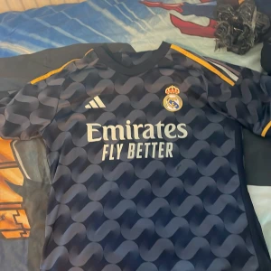 Real Madrid fotbollströja Adidas M - Snygg Real Madrid fotbollströja från Adidas i marinblått med vågigt mönster och gula detaljer på axlarna. Broderat klubbmärke på bröstet och tryckt sponsorlogga. Slim fit-modell i lätt och ventilerande polyester, perfekt för match eller träning.