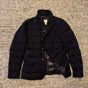 Moncler jacka  - Säljer en navy quiltad dunjacka från Moncler med klassisk kavajkrage och både dragkedja och knappar framtill. Jackan har flera fickor och en stilren, mörk look. Perfekt för dig som vill ha en varm och snygg jacka med exklusiv känsla. Notera skadan vid kragen som har fixat snyggt