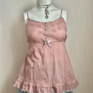 Rosa linnetopp med spets och rosett - Pink coquette-style top featuring ruffles, lace trim, and a charming bow. Please note: one button is missing. Tag says L but it fits more like medium so please check out the measurements 