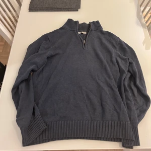 Blå stickad half zip-tröja Dressmann - Mysig blå stickad tröja från Dressmann i storlek M. Tröjan har en ribbad hög krage och half zip-dragkedja framtill. Perfekt för lager-på-lager och chillare dagar. Långa ärmar och bred ribbad mudd vid ärmslut och nederkant.