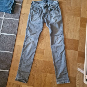 Replay Anbass grå jeans, storlek 29 - Snygga grå jeans från Replay, modell Anbass. De har en slim passform med klassiska fem fickor och Replay-logga på fickan. Jeansen är i stretchigt jeanstyg och har en modern, smal siluett som sitter riktigt bra. Finns ett litet hål