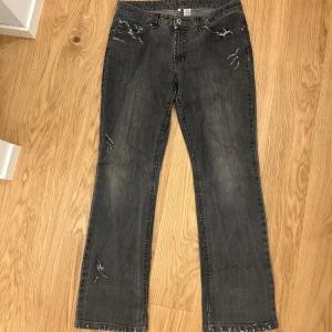 Grå slitna bootcut jeans med detaljer - Grå jeans med bootcut passform och slitna detaljer på både fram- och baksida. Råa kanter nertill och dekorativa sömmar på bakfickorna. Jeansen har en avslappnad vibe och är tillverkade i klassiskt denimtyg. Det är lite stora för mig och jag har normalt sett storlek 32, dessa skulle funka 32-35 skulle jag gissa på