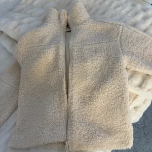 Beige teddyjacka XXS - Supermjuk och fluffig teddyjacka i beige med hel dragkedja framtill. Jackan har hög krage och långa ärmar, perfekt för kyliga dagar. Den har en oversized look och är tillverkad i mysigt fleece-material. Helt ny aldrig använd, från Gina Tricot!