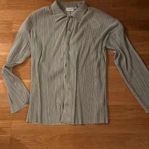 Ljusgrön plisserad skjorta från Kappahl - Snygg ljusgrön skjorta från Kappahl med plisserat tyg och klassisk krage. Skjortan har långa ärmar och knäppning framtill med matchande knappar. Den är oversize och funkar för både xs och s. Perfekt för dig som gillar stilrena och trendiga plagg med en twist.