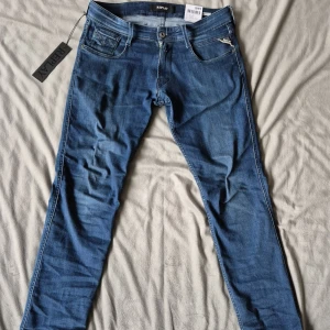 Blå Replay Anbass jeans slim fit - Snygga blå Replay Anbass jeans med klassisk femficksdesign och diskreta slitningar. Jeansen har slim passform, normal midja och är tillverkade i stretchig bomullsdenim för extra komfort. Perfekta för dig som gillar en modern och stilren look.