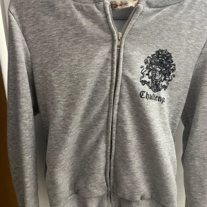 Grå hoodie med leopardfoder och tryck - Grå hoodie med dragkedja från Challenge, har ett svart tryck på bröstet och ett supermjukt leopardmönstrat foder i huvan. Perfekt för dig som vill ha något unikt och mysigt. Långärmad och med ribbade muddar.