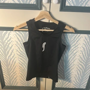 Svart sportig linnetopp RS Sports - Svart linnetopp från RS Sports med vitt logotryck framtill. Toppen är ärmlös och har en sportig, tight passform som passar perfekt för träning. Tillverkad i ett syntetmaterial som andas och ger bra rörelsefrihet.