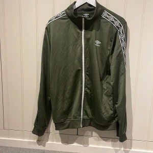 Grön track jacket från Umbro - Snygg grön track jacket från Umbro med vita detaljer och dragkedja framtill. Jackan har ribbade muddar, hög krage och vita mönstrade band längs ärmarna. Säljer då jag inte är något fan av den stora kragen. Hör av dig om du har frågor!