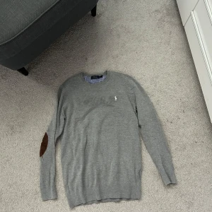 Grå tröja från Polo Ralph Lauren - Snygg grå tröja från Polo Ralph Lauren med rund halsringning och klassisk logga på bröstet. Tröjan har långa ärmar och ribbade muddar samt en brun detalj på ena armbågen. Perfekt för lager-på-lager-stil.