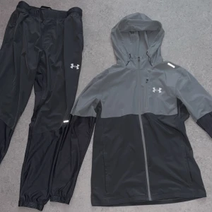 Under Armour vindjacka grå/svart M - Snygg vindjacka från Under Armour i tvåfärgad grå och svart design. Jackan har huva, hel dragkedja framtill och reflekterande detaljer. Perfekt för löpning eller utomhusträning. Tillverkad i lätt syntetmaterial för maximal rörelsefrihet.