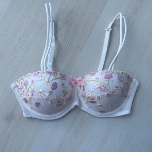 Vit blommig bandeau topp med broderi - Supersöt vit bh med broderade blommor i rosa, gult och grönt. Dubbla axelband och en liten rosett framtill. Kuporna är vadderade. Storlek S men motsvarar 70B. Aldrig använd då den är för liten🩷