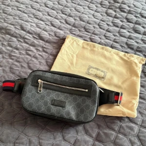 Gucci svart axelväska med monogram - Snygg svart axelväska från Gucci med klassiskt GG-monogram, silverfärgad dragkedja framtill och justerbart band med röd-blå rand. Väskan är i syntetmaterial med detaljer i skinn och har en stilren, rektangulär form. Perfekt för att bära det viktigaste.