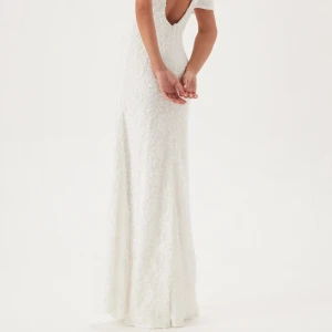 Vit långklänning med 3D-blommor - Superfin vit långklänning med kort ärm och v-ringning. Klänningen har 3D-blommor över hela tyget och en öppen rygg som ger en elegant vibe. Perfekt för speciella tillfällen när du vill sticka ut lite extra.