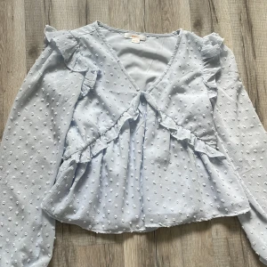 Ljusblå volangblus från Gina Tricot - Supersöt ljusblå blus från Gina Tricot med volangdetaljer och prickig struktur. Blusen har v-ringning, långa ärmar och är tillverkad i ett lätt och luftigt material. Perfekt för vår och sommar när du vill ha något extra fint.