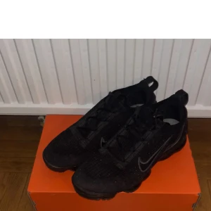 Svarta Nike Vapormax sneakers - Säljer ett par helsvarta Nike Vapormax sneakers med stickad ovandel och klassisk Swoosh på sidan. Skorna har snörning och den ikoniska genomskinliga sulan som ger en futuristisk vibe. Perfekta för dig som gillar sportig och stilren look.