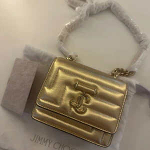 Guldig quiltad handväska från Jimmy Choo - Supersnygg liten handväska från Jimmy Choo i metallicguldigt skinn med quiltad design och JC-logga framtill. Väskan har kedjerem och magnetlås, perfekt för att lyfta vilken outfit som helst. Kommer med dustbag och äkthetskort.