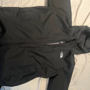 Svart vindjacka från The North Face - Svart vindjacka från The North Face med huva och dragkedja framtill.  Jackan är nästan helt ny och använd bara ett fåtal gånger. Jackan har meshfoder på insidan och logga tryckt på både bröstet och ryggen. Perfekt för blåsiga dagar och enkel att matcha med allt. Snygg och stilren design.