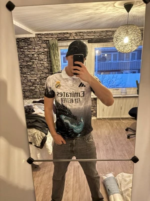 Real Madrid x Adidas draktröja - Säljer en unik Real Madrid t-shirt från Adidas med fet drakdesign i svart, vitt och turkost. Tröjan har korta ärmar, klassisk krage och tryck med 'Emirates Fly Better' på bröstet. Perfekt för dig som vill sticka ut på läktaren eller i stan.