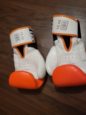 Vita och orange boxningshandskar från Yokkao - Säljer ett par boxningshandskar från Yokkao i storlek 12 oz. Handskarna är vita med orange detaljer och har svart insida vid handleden. Tillverkade i skinn med kardborreband för enkel justering. Perfekta för träning och sparring.