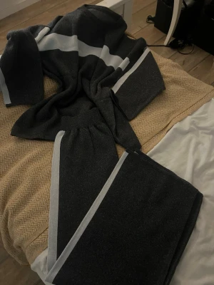 Mörkgrå stickad tröja med vit rand - Säljer en mörkgrå stickad tröja med bred vit rand över bröstet och längs ärmarna. Tröjan har en rund hals och långa ärmar med vit kant. Perfekt för dig som gillar enkel och stilren design med en sportig touch.