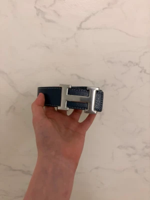 Blått bälte  - Säljer ett stilrent blått bälte med ett stort H-format spänne i silverfärg. Bältet har vit kontrastsöm längs kanterna och är gjort i ett slätt material som ger en clean look. Perfekt accessoar för att lyfta jeans eller byxor.