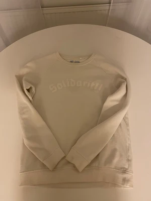 Beige Vit sweatshirt  - Säljer en vit sweatshirt från 157 DOG med trycket 'Solidarity' ton-i-ton på bröstet. Tröjan har rund hals, långa ärmar och ribbade muddar vid ärmslut och nederkant. Perfekt för en chill och stilren look.