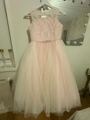 Ljusrosa långklänning med spets( flickkläder) - Superfin flickklänning ljusrosa långklänning med broderad spets upptill och fluffig tyllkjol. Klänningen har ärmlös design, knappar i ryggen och ett bälte med glittriga detaljer i midjan. Perfekt för speciella tillfällen och ger en riktigt drömmig känsla.