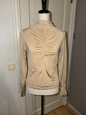 Beige långärmad polotopp från Indiska - Säljer en beige långärmad polotopp från Indiska i mjukt, glansigt material. Toppen har snygg rynkning framtill längs mitten och en hög krage som ger en clean look. Perfekt att styla med jeans eller kjol för en enkel men chic outfit.