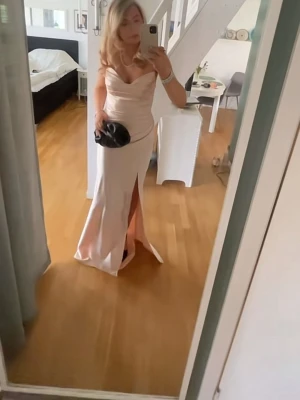 Champagnefärgad långklänning med slits - Elegant långklänning i champagnefärgad satin med figurnära passform. Klänningen har v-ringad omlottliv, smala axelband och en hög slits framtill som ger en snygg siluett. Perfekt för bal eller festligare tillfällen.