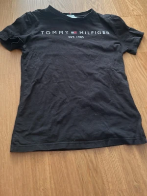 Svart Tommy Hilfiger t-shirt - Snygg svart t-shirt från Tommy Hilfiger med klassisk logga och texten 'EST. 1985' på bröstet. Tillverkad i ekologisk bomull för extra skön känsla. Enkel och stilren modell med korta ärmar och rund hals.