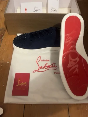 Mörkblå Christian Louboutin sneakers - Säljer ett par mörkblå sneakers från Christian Louboutin med klassisk röd sula och coola nitar över hela tån. Skorna har snörning och är gjorda i mocka med vita kanter runt sulan. Perfekta för dig som vill sticka ut med exklusiv design. Hör av er vid frågor✅
