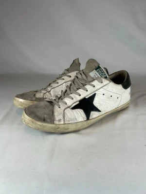 Golden Goose vita sneakers med svart stjärna - (Nypris 4900kr)tjena säljer ett par äkta Golden Goose sneakers i vitt skinn med grå mockadetaljer på tån och grå snörning. Sidan har en stor svart stjärna och hälen är mörkgrön. Platt sula med vintage-look och GGDB-märke på plösen. Perfekt för dig som gillar streetstyle och unika detaljer.(Size 44-45)