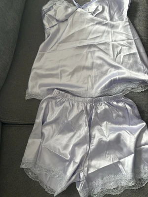 Ljuslila pyjamas med spetsdetaljer - Säljer en ljuslila pyjamas i glansigt sidenmaterial med tunna axelband och spetskant nedtill. Pyjamasen har en feminin v-ringning och känns riktigt mjuk och lyxig. Perfekt för dig som gillar en elegant och chill vibe.