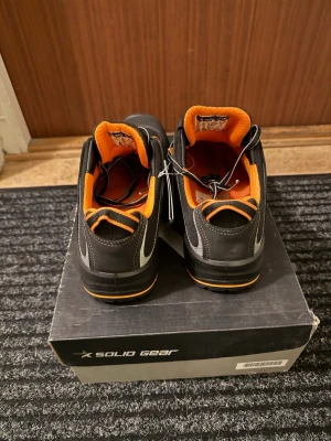 Solid Gear jobbskor  - Säljer ett par stabila jobbskor/syddsskor i svart med orange detaljer. Skorna är gjorda för arbete inom t.ex. lager, industri, verkstad eller serviceyrken där man står och går mycket.