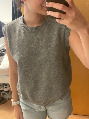 Grå stickad väst från Zara - Säljer en enkel och snygg grå stickad väst från Zara. Modellen är rak med ribbade kanter vid halsringning, ärmhål och nederkant. Perfekt att slänga över en skjorta eller t-shirt för en chill look. Passar även S-M