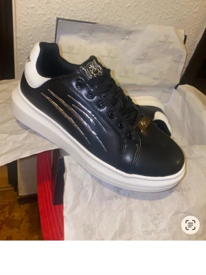 Svarta Plein Sport sneakers med silvriga detaljer - Snygga svarta sneakers från Plein Sport med vita sulor och häldetaljer. Skorna har silvriga klomärken på sidan, snörning och en cool logga på tungan. Tillverkade i syntetmaterial med en chunky platt sula och rund tå. Perfekta för dig som vill sticka ut.