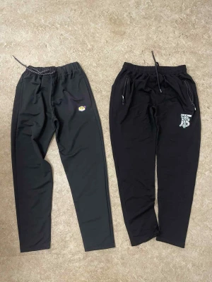 Svarta mjukisbyxor med tryck - Två par svarta mjukisbyxor, ena med Tn-logga och den andra med vit TB-logga. Båda har dragsko i midjan och smal passform. Ena paret har dragkedjor på fickorna. Perfekta för chill eller träning.