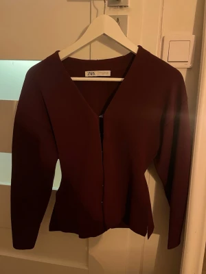 Vinröd kofta med v-ringning från Zara - Snygg vinröd kofta från Zara med v-ringad hals och dragkedja framtill. Modellen har lång ärm och en figurnära passform som ger en stilren siluett. Perfekt att matcha med jeans eller kjol för en trendig look.