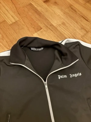Svart track jacket från Palm Angels - Ny äkta Palm Anglès kofta! Säljer en svart track jacket från Palm Angels med vita detaljer längs ärmarna och vit dragkedja. Jackan har hög krage, två fickor med dragkedja och Palm Angels-logga på bröstet. Perfekt för en sportig och trendig look.