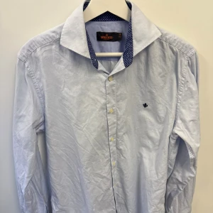 Ljusblå skjorta från Morris - Snygg ljusblå skjorta från Morris med diskret rutmönster och mörkblå prickig insida på kragen. Klassisk passform med knappar framtill och broderad logga på bröstet. Perfekt till jeans eller chinos för en clean look.