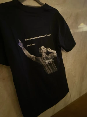 Zlatan T-Shirt - Lyft din stil med vår svarta premium T-shirt med ett svartvitt tryck av Zlatan på ryggen. En stilren och kraftfull design som hyllar en av världens största fotbollsstjärnor – perfekt för både vardag och streetwear-look. T-shirten är tillverkad i mjuk och slitstark bomull som ger hög komfort hela dagen. Den minimalistiska framsidan kombineras med ett iögonfallande motiv på ryggen, vilket skapar en modern och trendig kontrast  