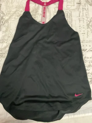 Svart linnetopp från Nike - Säljer en svart linnetopp från Nike med rosa detaljer och texten 'JUST DO IT.' på ryggen. Toppen har tunna axelband och en sportig look, perfekt för träning. Materialet är mjukt och följsamt syntet som andas bra.
