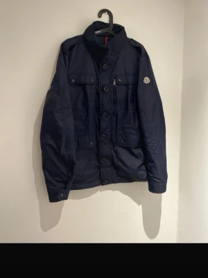 Mörkblå Feildjacka från Moncler - Snygg mörkblå Feildjacka från Moncler med klassisk krage där loggan är broderad. Jackan har flera fickor med knappar framtill och detaljerade manschetter med tryckknappar. Perfekt för dig som gillar stilrena och funktionella plagg. Använd ca 3 gånger och är i nyskick. Vid snabb affär kan priset diskuteras.